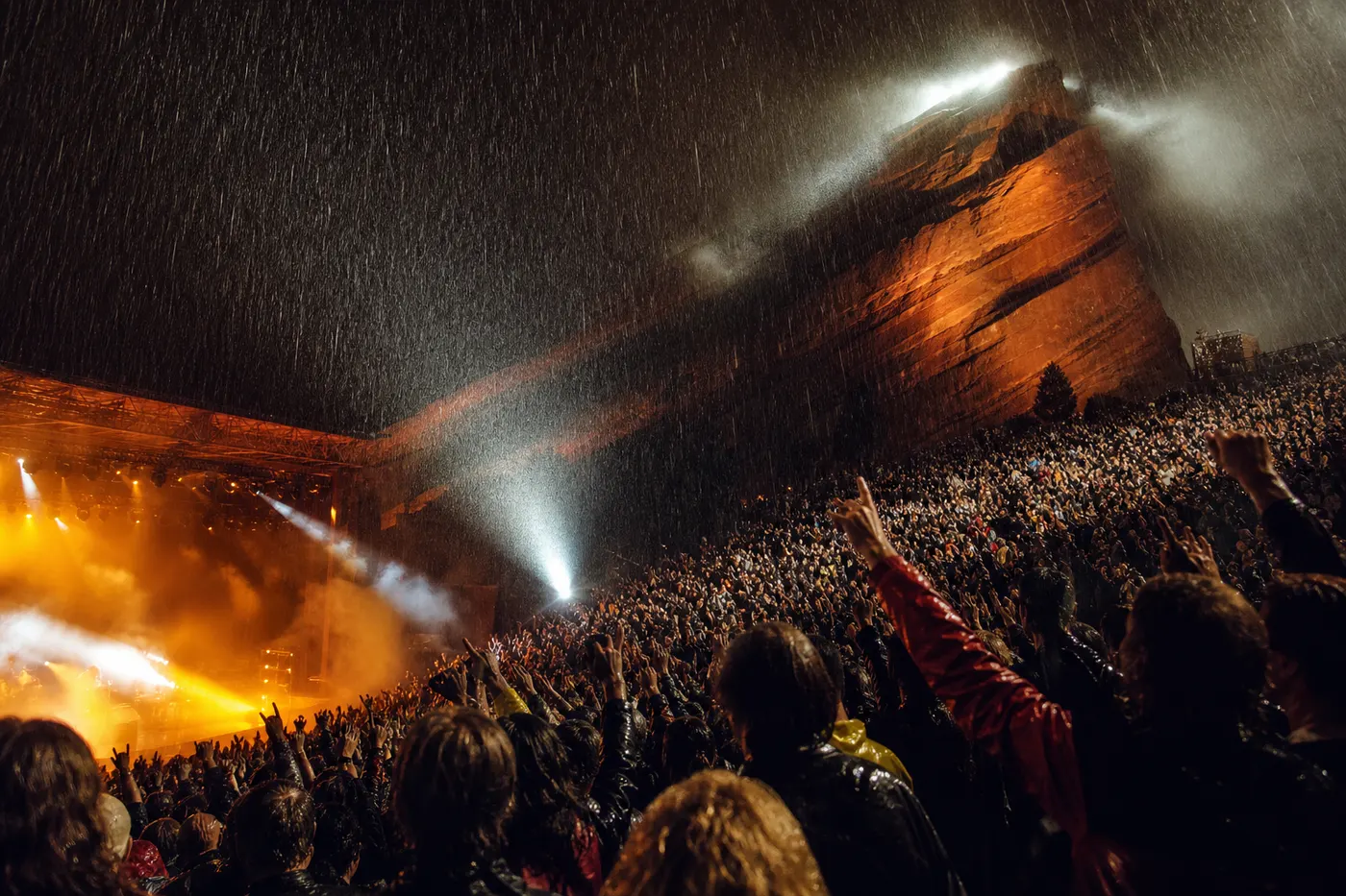 Dramatic concert at Red Rocks in the rain — crowd with arms raised, massive red rock formation illuminated by stage lights, rain pouring through spotlights