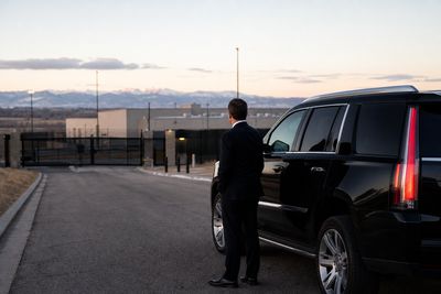 Chauffeur standing beside a black SUV on an approach road to a secure Colorado facility at dawn